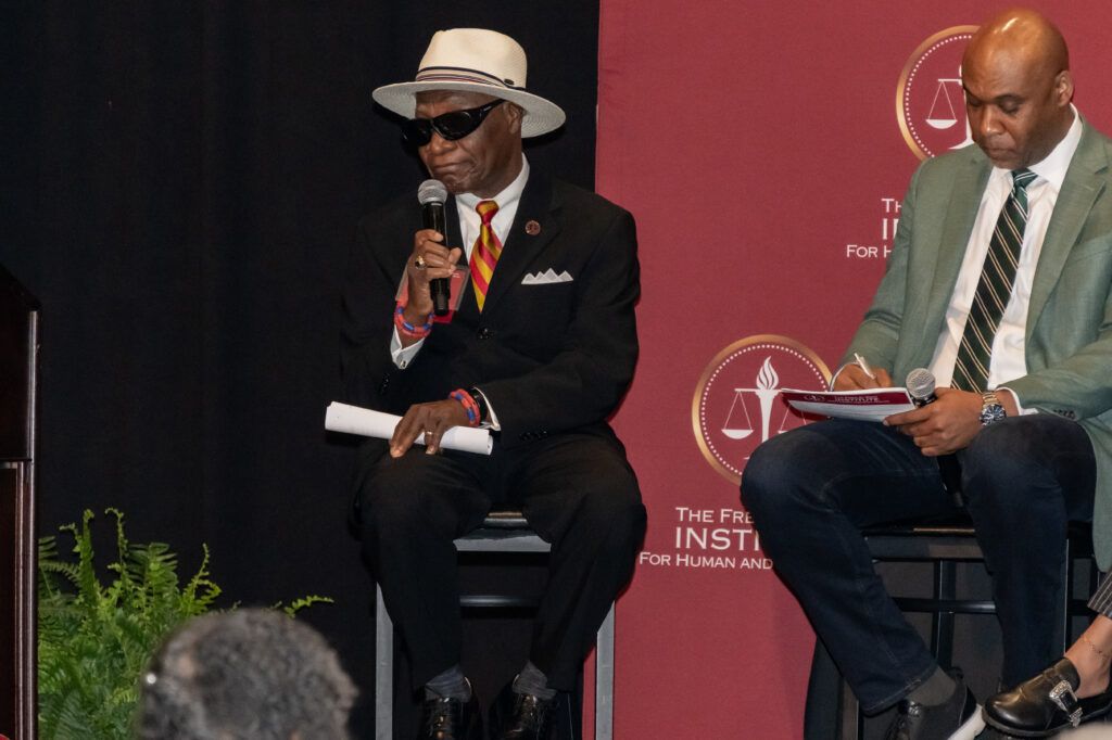 Two men sit on stage in front of a maroon backdrop featuring an emblem. The man on the left wears sunglasses, a hat, and holds a microphone. The man on the right is reading notes. Both are dressed in formal attire. A podium and potted plant are visible.