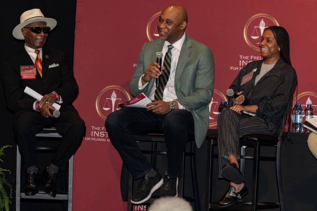 Three people are seated on a stage against a maroon backdrop with logos. A man in a hat and sunglasses sits on the left, holding papers. The man in the center speaks into a microphone. A woman on the right smiles, also holding a microphone.