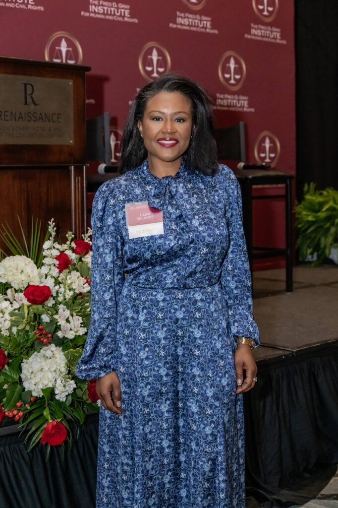 A woman in a blue patterned dress stands in front of a stage with a maroon backdrop featuring several emblems. She is wearing a name badge and smiling. A large floral arrangement is visible to her left.