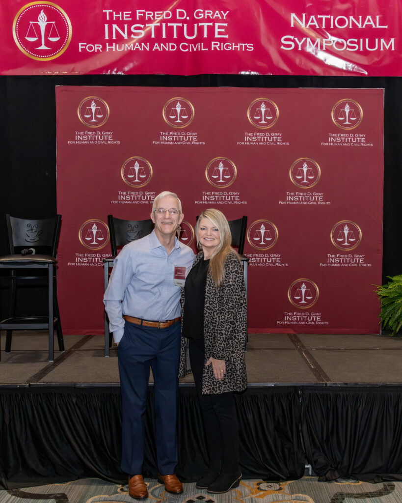 A man and a woman stand smiling in front of a backdrop with "The Fred D. Gray Institute for Human and Civil Rights" logo. They are on a stage with two empty chairs to the left. A banner above reads "National Symposium.