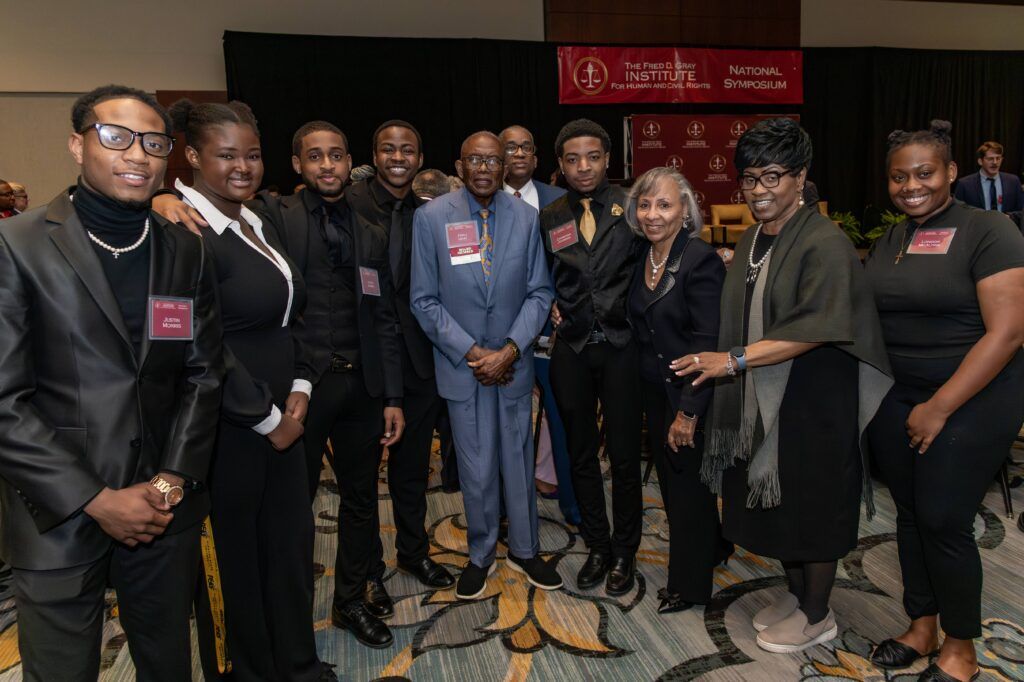 A group of people in formal attire stand together for a photo at an event. A banner in the background reads “The Fred Davis Institute National Symposium.” They are smiling and posing in a well-lit room with patterned carpet.
