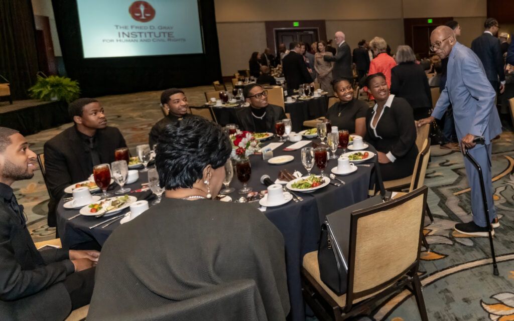 A group of people dressed in formal attire are sitting at a round table with plates of salad and drinks. A man with glasses and a cane stands beside the table. A projection on a screen reads "The Fred D. Gray Institute for Human and Civil Rights.