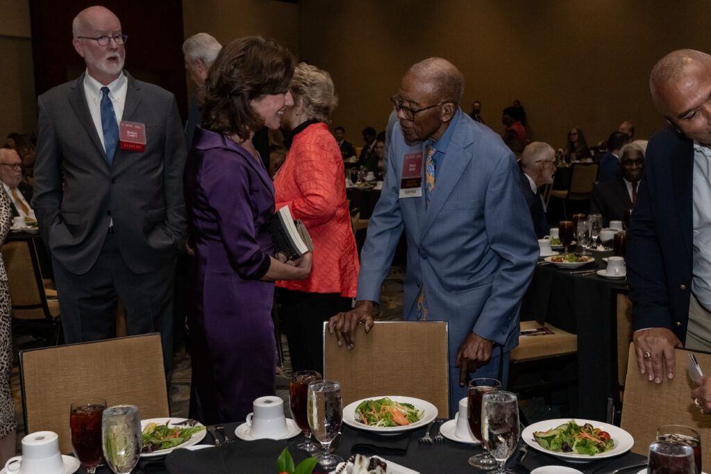 A group of people in formal attire are gathered in a large room with dining tables. Two individuals in the foreground are conversing, surrounded by plates of salad, glasses, and place settings.
