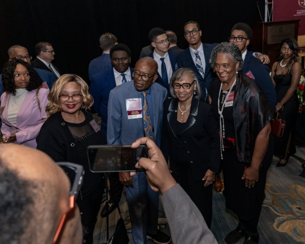 A group of people, dressed in formal attire, gather for a photo at an indoor event. Someone in the foreground holds up a phone to take a picture. A podium and banner are partially visible in the background.