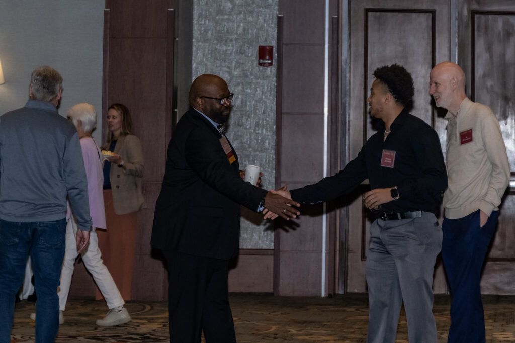 Two men shaking hands in a conference room, both wearing name tags. Three other people are walking in the background, holding drinks. The room has a carpeted floor and wooden paneled walls.