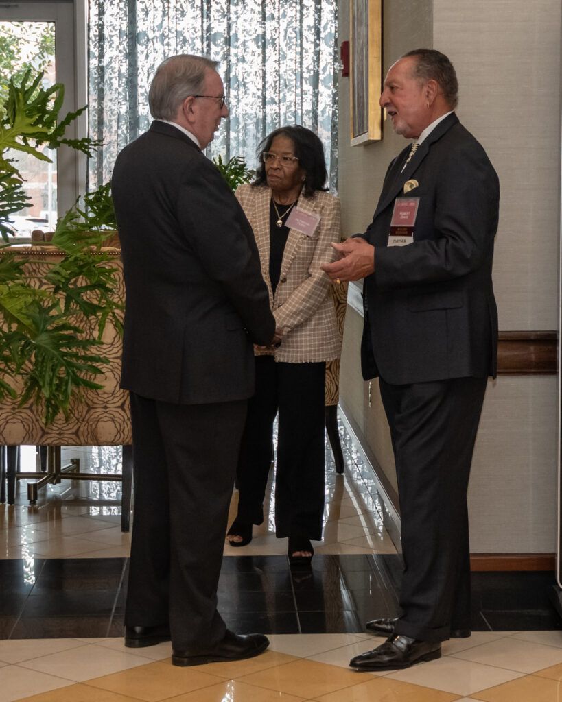 Three people in formal attire are talking in a well-lit room with green plants around. Two men in suits are standing and engaged in conversation, while a woman in a checkered blazer observes. A large window with curtains is in the background.