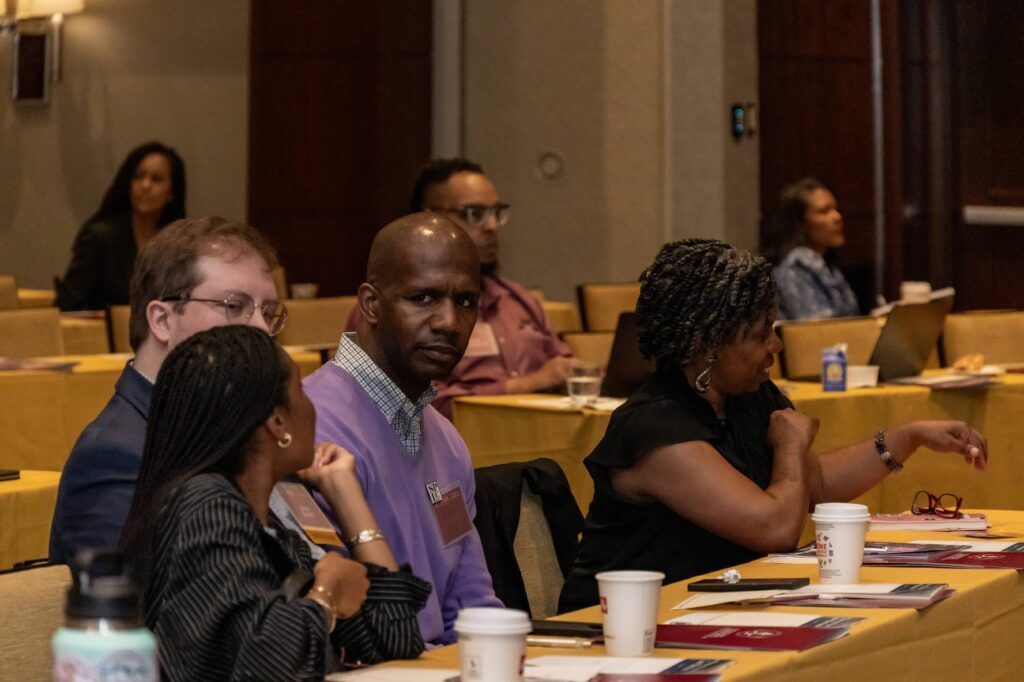 A group of people seated at a conference room with yellow tablecloths. Some have cups and notebooks in front of them. A man in a purple sweater is facing forward while others are looking in the same direction or conversing among themselves.