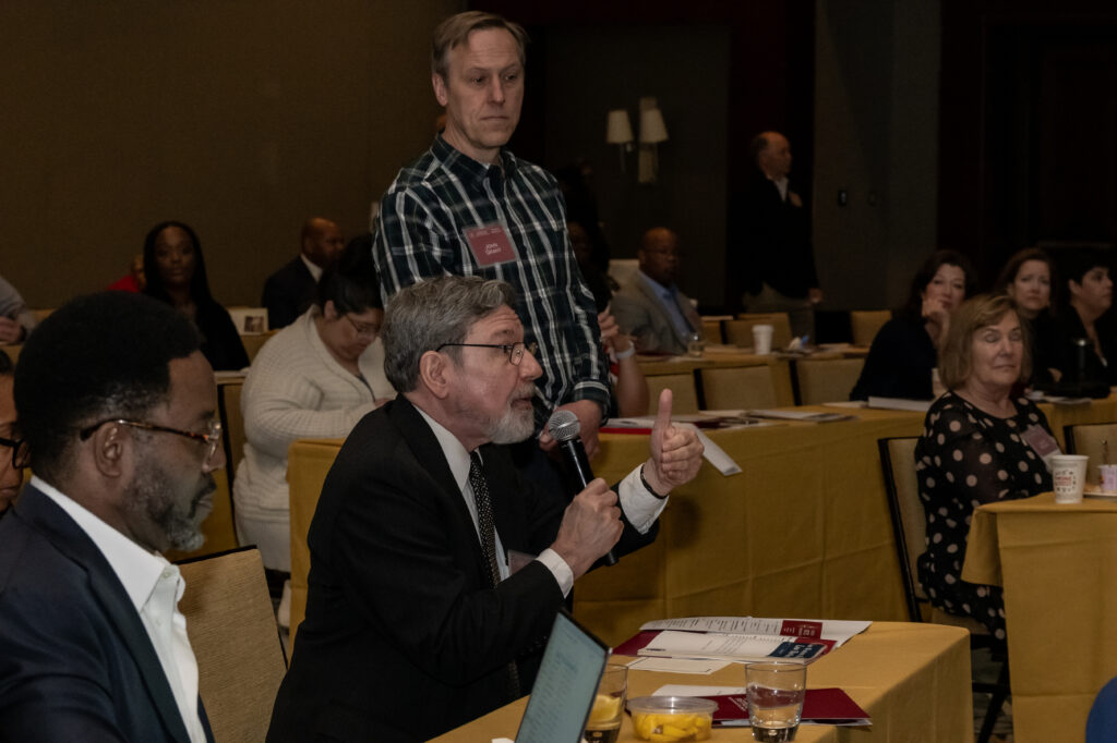 A person with a microphone gestures while speaking at an indoor event, surrounded by attendees seated at tables with yellow tablecloths. A standing attendee listens attentively. Several people and documents are visible in the background.